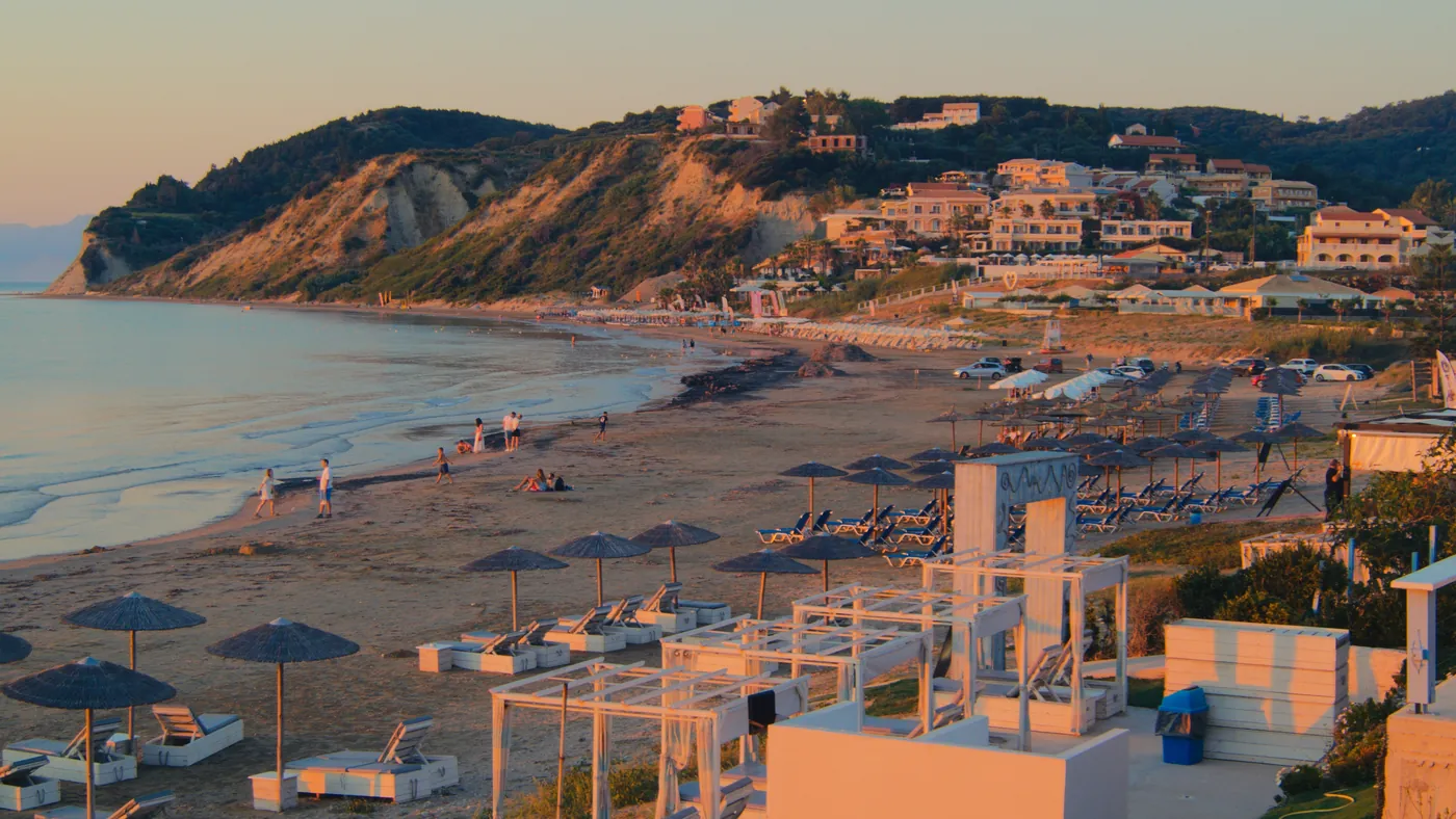 A beach in Corfu at golden hour, with sun umbrellas on the sand and hillside villas behind